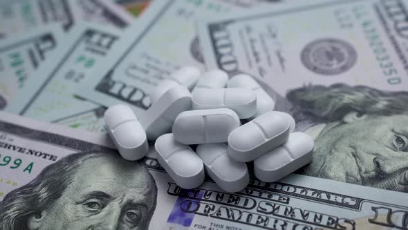 A pile of white pills rests on the one hundred dollar bills scattered on the table alt