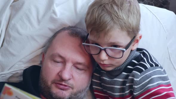 Father Reads a Book to His Son Solve Riddles in Bed Closeup