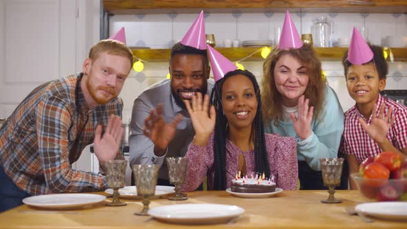 Portrait of Multiethnic Family Sitting at Table and Waving Hands at Camera Having Birthday Party alt