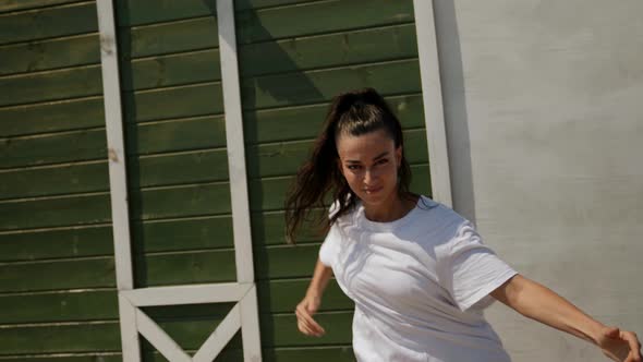 a Brunette in a Big White Tshirt Dances Against a Green Wall on the Street on a Sunny Day alt