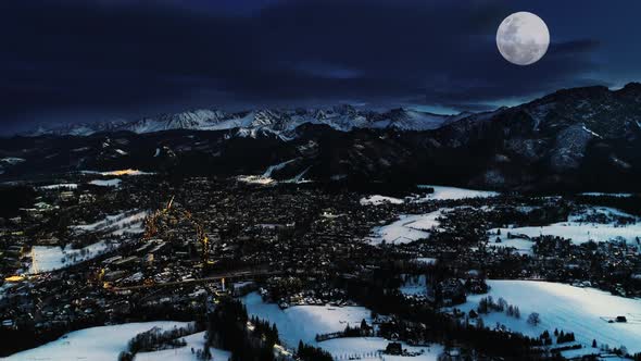 Mountains at winter night, aerial scenic view. Illuminated city covered with snow, magical winter