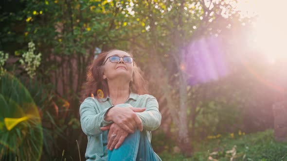 A Mature Adult Woman in Glasses Sits in a Country House's Garden alt