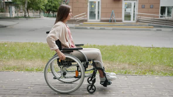 Side View of Beautiful Disabled Woman Moving in Wheelchair Along City Street alt