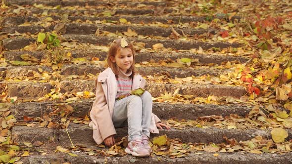 a Cute Little Girl Sitting on a Staircase with Fallen Foliage in an Autumn Park alt