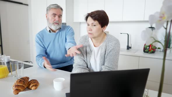 Brunette Middle Aged Woman Talking with Worried Greyhaired Senior Man Sitting in Kitchen with Laptop alt