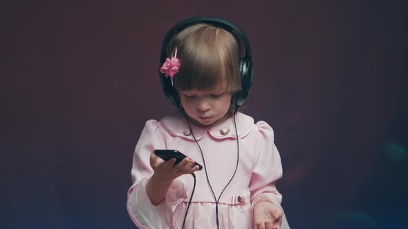 Little Girl in Big Headphones and Vintage Dress is Listening to Music on Phone alt