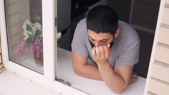 A young man goes to the window in a medical mask and removes it to breathe fresh air. alt