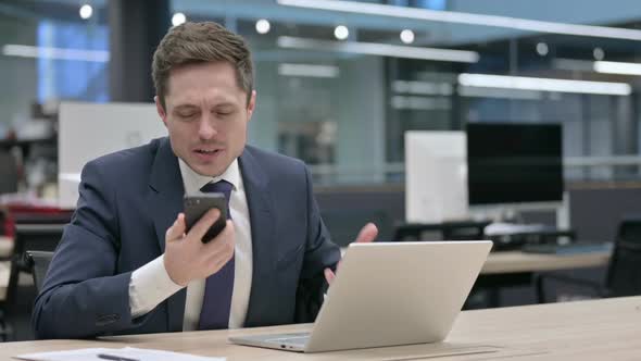 Angry Businessman Talking on Smartphone While Using Laptop in Office alt