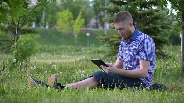 a Man in an Open-air Park Sits on the Grass and Uses a Tablet. Against Quarantine and Home alt