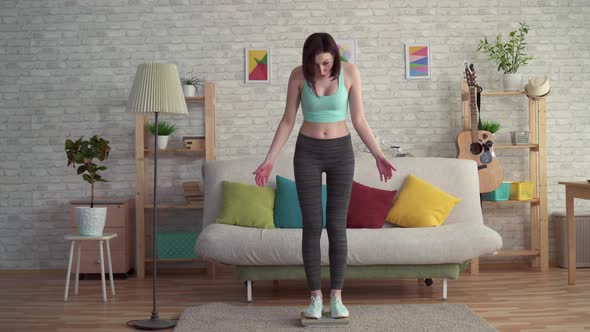 Expressive Cheerful Young Woman in Sportswear Measures Her Weight on the Floor Scales alt