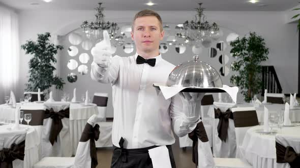 A Male Waiter in a White Shirt in the Restaurant Hall Shows a Raised Thumbs Up and Down alt