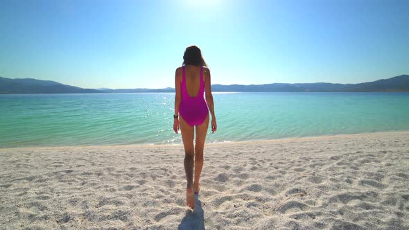 Sexy Female Model in Pink Swimsuit Walking on the White Beach of the Clear Tropical Turquoise Sea alt