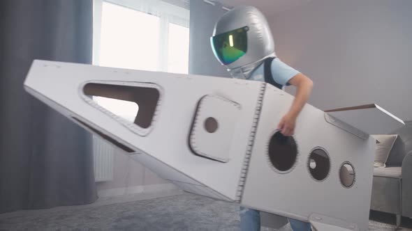 Kid Boy in an Astronaut Costume Plays with a Cardboard Space Shuttle Runs Around the Room Slow alt