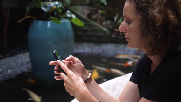 Side View of a Young Woman Browsing on Her Phone While Sitting Next to the Pond with the Goldfishes alt
