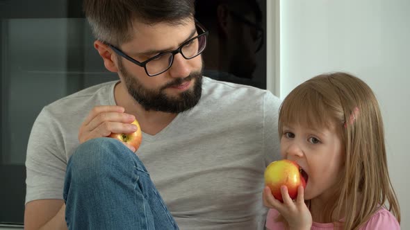 Happy Family Father and Daughter Sit on Floor in Kitchen at Home and Eat Apples
