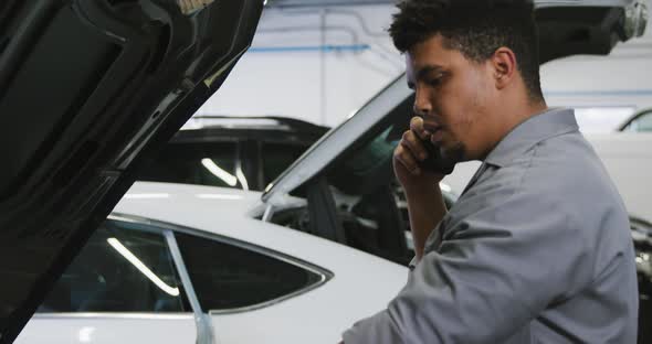African American male car mechanic looking at an open car engine and talking on a smartphone alt