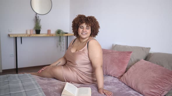 Beautiful Curvy Plus Size African Black Woman Afro Hair Lying on Bed in Silk Powder Pink Dress Cozy