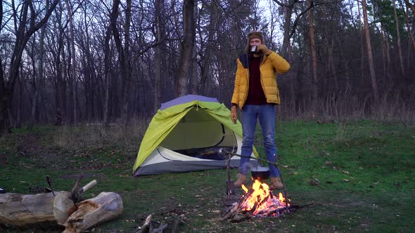A Man Drinks Tea Near a Campfire and a Tent in the Forest alt