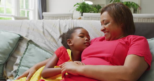 Happy african american grandmother and granddaughter hugging and talking on sofa alt