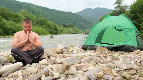 The Fat Man Practices Yoga Near a Mountain River Followed By a Green Tent alt