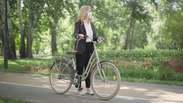 Wide Shot of Confident Caucasian Middle Aged Woman in Suit and Coronavirus Face Mask Sitting on Bike alt