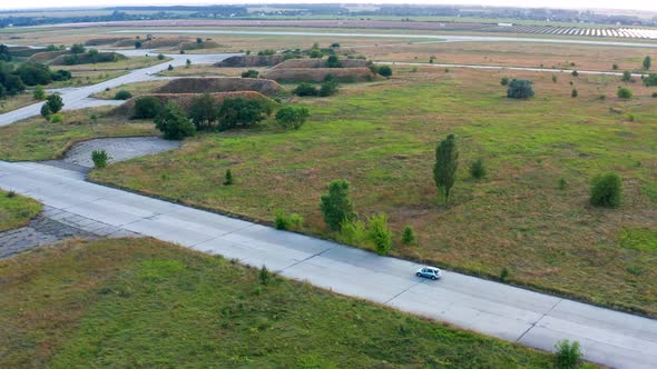 Flying over a car that is traveling on an airplane runway. Forest and grass on the sides. alt