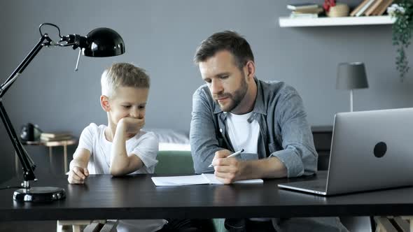Man Helping Young Boy In Room Doing Homework And Smiling, Studying, Sitting at A Table alt