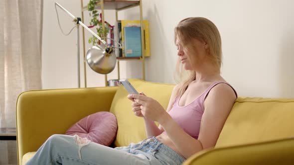 Blonde Sits on Yellow Sofa in Pink Top and Use Phone Typing Text alt