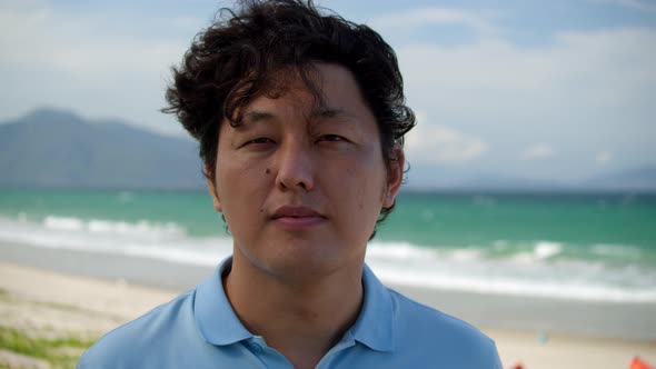 Portrait of a Middleaged Asian Man in a Blue Shirt Standing on the Beach By the Sea Against the alt