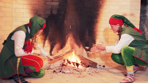 Elves in green fairytale costumes are sitting by a large fireplace with fire. alt