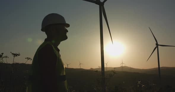 A Young Engineer Enters the Frame and Then Examines the Windmill in Front of Him at Sunset alt