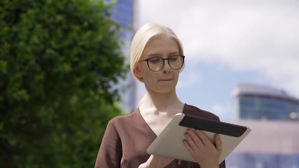 A Business Woman in a Formal Dress and Glasses Scrolls the Tablet in the Street