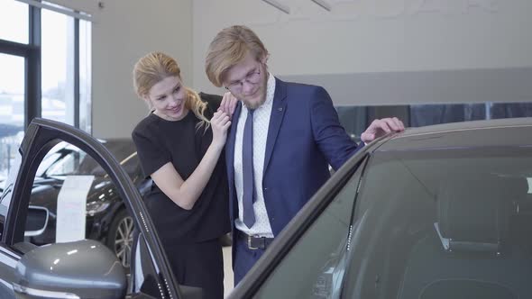 Joyful Man and Woman Just Bought Car in Modern Motor Show. Couple Hugging Near Automobile alt