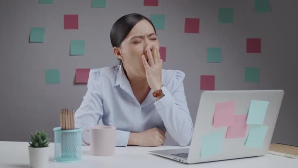 Asian woman working on a laptop at home office sleepy and yawning.