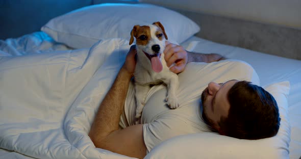 a Man Is Lying on a Sofa with White Bed Linen with His Dog. the Guy Holds the Animal on His Stomach alt