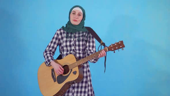 Puzzled Young Muslim Woman Clumsily Plays Guitar on Blue Background alt