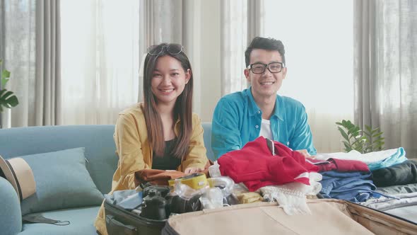 Happy Asian Couple Travelers Packing Clothes In A Suitcase And Looking At Camera alt