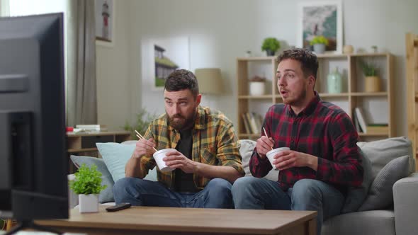 Two Young Cute Men Caucasian Eating a Chinese Noodle Sitting in a Living Room on a Couch and Watch alt
