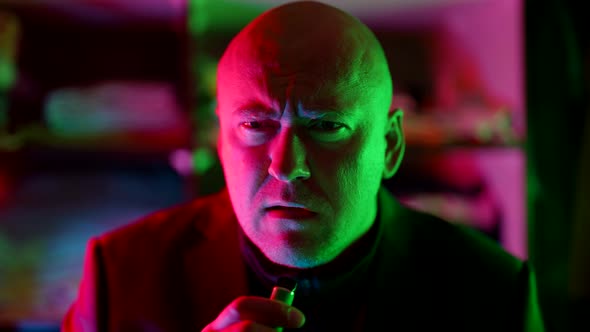Middleaged Bald Man is Smoking Ecigarette in Nightclub Male Portrait in Flashing Neon Lights alt