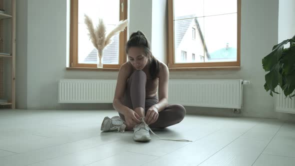 Young Girl in Sportswear Sits on Floor on Knees Untying Shoelaces on Sneakers