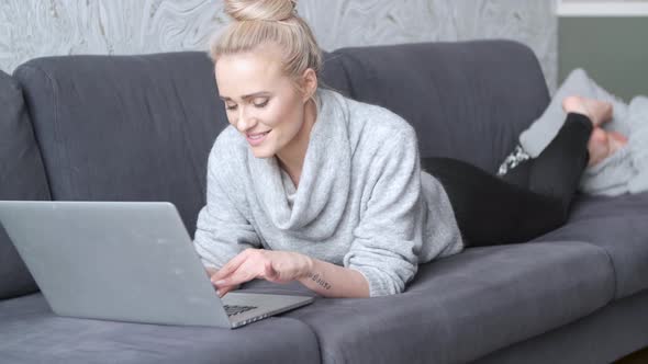 Happy Blond Woman Lying Prone on Sofa and Working on Laptop Computer alt