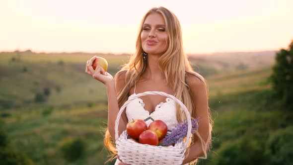 Beautiful sexy blonde girl in white dress posing in a field at sunset with a basket of fruit alt