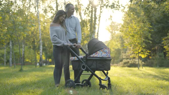 Wide Shot of Happy Smiling Husband and Wife Standing with Baby Stroller in Sunny Park and Talking alt