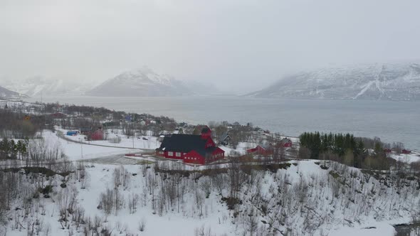 Aerial drone orbits around Kåfjord Church in Norway. Snow covered coastline, with mountainous backgr alt