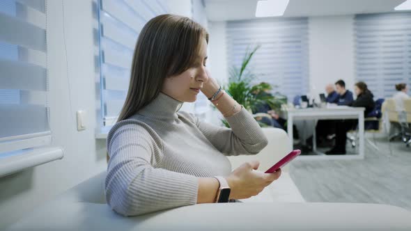 Closeup of a Modern Girl Uses a Mobile Phone on the Couch in the Office Scrolls Types Text Surfs the alt