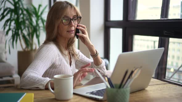 Businesswoman Talking on Phone and Sitting at Table with Laptop in Home Office alt