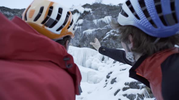 Two men point and discuss a climbing route for ice climbing on a frozen waterfall in the mountains. alt