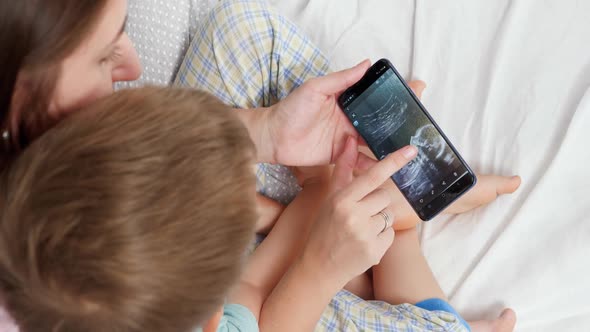 Closeup of Young Woman Showing Baby Ultrasound Image To Her Little Son on Smartphone alt