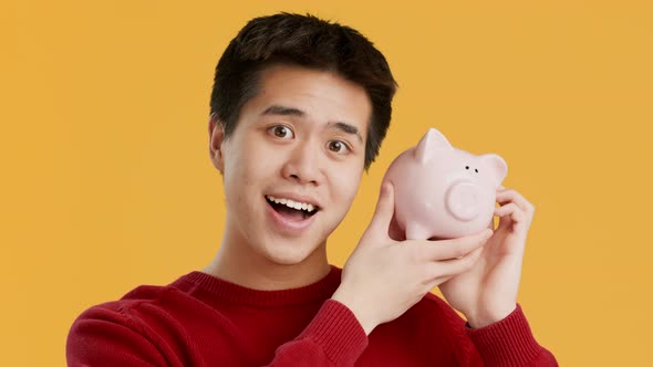 Rich Asian Man Shaking Piggybank Full Of Money Yellow Background alt