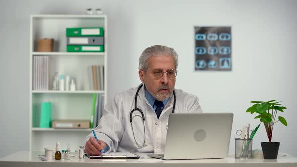 Aged Male Doctor Reviews the Patient's Analyzes on a Laptop and Makes Notes in His Medical History alt